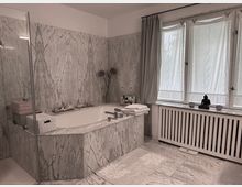 Modernes Badezimmer mit einer Badewanne, die von marmorierten Wänden und Fliesen umgeben ist. Links befindet sich eine Glasduschkabine, während rechts große Fenster mit halbtransparenten Vorhängen Licht einlassen; auf der Fensterbank steht eine kleine Buddha-Statue.