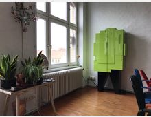 Ein helles Zimmer mit einem großen Fenster, das Licht hereinlässt und einen Blick auf rote Ziegeldächer bietet. Links stehen auf einem kleinen Tisch Zimmerpflanzen, rechts befindet sich ein auffälliger grüner, geometrisch gestalteter Schrank vor einer weißen Wand; Holzboden und bunte Stühle runden den Raum ab.