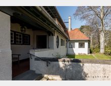 Einfamilienhaus im traditionellen Stil mit weißer Putzfassade, rotem Ziegeldach und Sprossenfenstern, umgeben von einem Garten mit Rasen und Bäumen. Im Vordergrund ein überdachter Eingangsbereich mit Geländer und einer Außenwand aus Beton.
