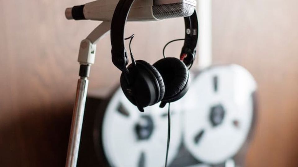 Mikrofon an einem Stativ mit darüberhängenden Kopfhörern; im unscharfen Hintergrund steht ein Tonbandgerät mit großen Spulen vor einer Wand.