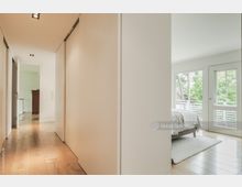 Flur eines modernen Wohnhauses mit hellen Wänden und Holzboden, der in ein Schlafzimmer mit großen Fenstern und Blick ins Grüne führt. Rechts befindet sich ein Bett und links ein geöffneter Durchgang zu einem weiteren Raum.