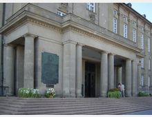 Fassade eines repräsentativen Gebäudes aus Naturstein mit einer Säulenfront und einem Treppenaufgang. Neben dem Eingang befindet sich eine Gedenktafel, davor sind Blumenkränze und Pflanzen platziert.