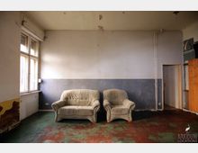 Ein einfacher, leicht heruntergekommener Raum mit einer beigen Couch und einem passenden Sessel vor einer zweifarbigen Wand (oben weiß, unten grau). Der Raum hat ein großes Fenster auf der linken Seite, eine beschädigte Tapete an der unteren linken Wand und einen abgenutzten rot-grünen Bodenbelag.