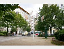 Städtischer Platz mit Bäumen, einer Metallskulptur mit farbigen Elementen in der Mitte und umliegenden historischen Wohngebäuden. Im Hintergrund sind parkende Fahrzeuge, Fahrräder und Sitzbänke zu sehen.