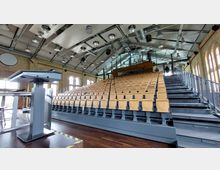 Ein moderner Hörsaal mit ansteigenden Sitzreihen aus hellem Holz, einer Glasrückwand und einem hohen Dach mit sichtbaren Metallstreben und Beleuchtung. Links im Vordergrund steht ein Rednerpult, während die Backsteinwände mit großen, bogenförmigen Fenstern gestaltet sind.