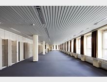 Großer leerer Innenraum mit Teppichboden, heller Deckenverkleidung aus schmalen Metallpaneelen und langen Fensterreihen mit braunen Vorhängen auf der rechten Seite. Linkerhand befinden sich offene Schrankelemente, die entlang der Wand verlaufen.