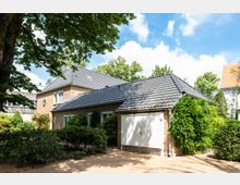 Zweistöckiges Einfamilienhaus aus Backstein mit grauem Satteldach, großen Fenstern und angrenzender Garage mit weißem Tor. Die Umgebung ist von Bäumen und Sträuchern umgeben, mit einem gepflasterten Weg im Vordergrund.