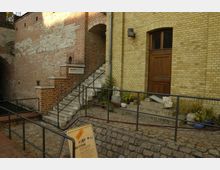 Backsteinarchitektur mit einer Treppe, die zu einem Schild mit der Aufschrift "Kunstbastion" führt. Daneben befindet sich eine Holz-Eingangstür, umgeben von Pflanzkübeln und einem Geländer aus Metall; im Vordergrund ein Informationsschild auf Kopfsteinpflaster.