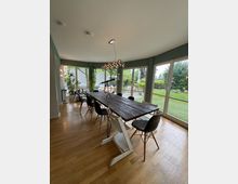 Esszimmer mit einem langen rustikalen Holztisch, umgeben von verschiedenen Stühlen, darunter ein Hochstuhl. Große Fensterfronten durchfluten den Raum mit Tageslicht und bieten Blick auf einen Garten mit Rasen und Pflanzen.