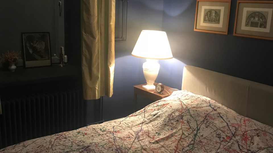Gemütliches Schlafzimmer mit dunkelblauen Wänden, einem Bett mit farbgesprenkelter Bettdecke und einem Nachttisch mit leuchtender Tischlampe. Links ein Fenster mit Vorhang und Fensterbank, rechts eine Wand mit mehreren gerahmten Bildern.