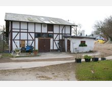 Fachwerkgebäude mit weiß verputzten Wänden und braunen Holzstreben, ergänzt durch einen kleinen Anbau mit Flachdach. Im Vordergrund befinden sich ein gepflasterter Hof, mehrere Blumentöpfe und ein kleiner Rasenbereich.