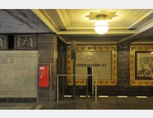 U-Bahn-Station mit der Beschilderung „Hohenzollernplatz“, gekennzeichnet durch beigefarbene und braune Fliesenwände, dekorative Reliefs und einen roten Notfallkasten. Die Decke ist mit einem runden Deckenleuchter und Gipsverzierungen gestaltet.