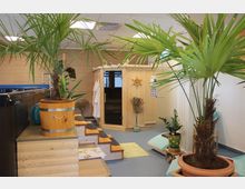 Wellnessraum mit einer Sauna aus hellem Holz in der Mitte, umgeben von tropischen Pflanzentöpfen. Der Raum verfügt über blaue Bodenfliesen, ein Spa-Pool am Rand und eine helle Einrichtung mit Handtüchern und Liegen.