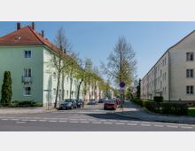 Straßenansicht eines Wohngebiets mit mehrstöckigen Häusern, links in mintgrün und rechts in beige. Die Straße ist von geparkten Autos und schmalen Bäumen gesäumt, im Hintergrund ist eine Kirche mit Turm erkennbar.