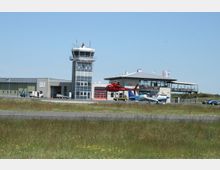 Kleiner Flughafen mit einem Kontrollturm, einem angrenzenden Hangar und einem zweistöckigen Gebäude mit Terrasse. Im Vordergrund starten oder landen ein roter Hubschrauber und ein kleines Flugzeug auf einer Asphaltpiste, umgeben von einer Wiese.