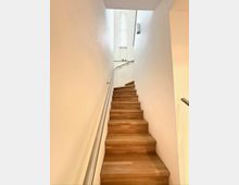 Schmales, modernes Treppenhaus mit Holzstufen und weißen Wänden, entlang einer Seite mit einem Metallhandlauf. Oben sorgt ein hohes, schmales Fenster für Tageslicht im Treppenaufgang.