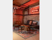 Gemütlicher Innenraum mit industriellen Elementen: Backsteinwände und freiliegende Metallrohre an der Decke, ein Lederfauteuil, eine Holzbank mit bunten Kissen, Holzwürfel als Couchtisch und Hocker mit Metallgestellen auf einem bunten Teppich; an der Wand ein leuchtendes Neon-Schild mit der Aufschrift "BERTA".