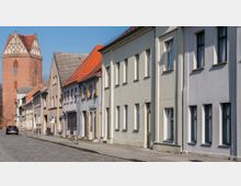 Straßenansicht einer historischen Altstadt mit schmalen, mehrstöckigen Wohnhäusern in hellen Farben und einem Kopfsteinpflaster. Im Hintergrund steht ein großer, roter Backsteinturm mit spitzem Dach und gotischen Elementen.