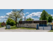 Eingezäuntes Industriegebäude mit flachem Dach und großen Fenstern, umgeben von Bäumen und einer Wiese. Im Vordergrund ein blaues Metalltor, links ein großer Baum und kleine ländliche Gebäude im Hintergrund.