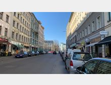 Straßenszene in einer urbanen Umgebung mit mehrstöckigen Wohn- und Geschäftshäusern auf beiden Seiten, flankiert von geparkten Autos. Am rechten Gehweg befinden sich verschiedene Geschäfte und Cafés, während im Hintergrund Bäume und ein modernes Gebäude sichtbar sind.