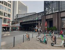 Bahnhofsvorplatz des Bahnhofs Friedrichstraße in Berlin mit umliegenden Gebäuden, darunter ein modernes Bürogebäude und ein Backsteingebäude. Vor dem Bahnhof stehen abgestellte E-Scooter, Fahrräder und Sitzgelegenheiten, während in der Umgebung Menschen laufen und Gespräche führen.