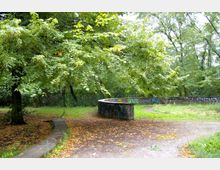 Parklandschaft mit dichtem grünen Baumbestand und einer geschwungenen, niedrigen Steinmauer, die teilweise mit Graffitis bedeckt ist. Der Boden ist teils mit Gras, teils mit Laub bedeckt, und ein geschwungener Weg führt durch die Szene.