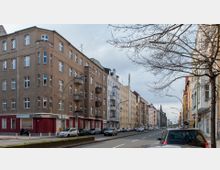 Straßenszene in einem städtischen Wohngebiet mit mehrstöckigen Putzbauten in verschiedenen Farben und klassischen Fassaden. Am Straßenrand parken Autos, während kahle Bäume und eine breite Fahrbahn die Umgebung prägen.