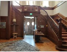 Holzvertäfelter Eingangsbereich mit einer geschwungenen Holztreppe, die zu einer oberen Etage führt, sowie einer Doppeltür mit runden Fenstern im Hintergrund. Der Raum ist mit Teppichboden und klassischen Möbeln wie einer Tischlampe und einem kleinen Hocker eingerichtet.