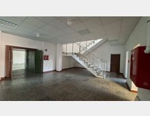 Geräumiges Treppenhaus in einem Gebäude mit gefliester Bodenfläche, weißer Rasterdecke und einer Treppe mit Metallgeländer. Links befindet sich ein offener Durchgang zu einem weiteren Raum, rechts sind eine Tür sowie ein roter Feuerlöschkasten und Feuerlöscher an der Wand zu sehen.