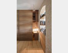 Innenraum mit einem kleinen Arbeitsplatz, der hinter einer Holzschiebetür verborgen ist. Rechts ist eine Ziegelwand mit Regalen voller Bücher, während der Boden aus hellem Holz besteht und eine minimalistische Hängelampe von der Decke hängt.