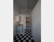 Schmale Küche mit schwarz-weiß kariertem Fliesenboden, weißen Wänden und schlichten Hängeschränken auf der linken Seite. Im Hintergrund führt eine Tür zu einem weiteren Raum mit ähnlichem Bodenmuster.