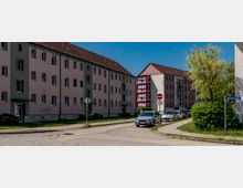Straßenszene in einem Wohngebiet mit mehrstöckigen, verputzten Mehrfamilienhäusern und roten Fensterrahmen sowie einem gläsernen Treppenhausanbau. Entlang der kurvigen Fahrbahn stehen geparkte Autos, daneben Gehwege, Grünflächen und ein Straßenschild „Joliot-Curie-Straße“.