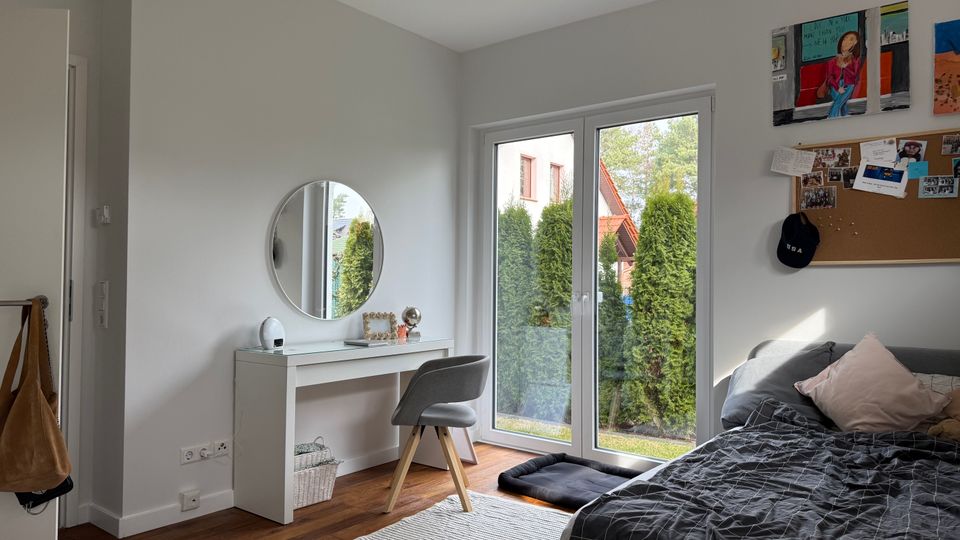 Bright modern bedroom with white walls, a low bed, and a small vanity desk with a round mirror and chair. Large glass patio doors open to a view of tall evergreen shrubs, and a corkboard with photos hangs on the wall.