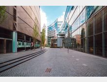 Moderner Stadtplatz mit gepflastertem Boden, flankiert von Gebäuden mit Glasfassaden und Metallstrukturen. Im Hintergrund ist ein rundes Gebäude mit weiteren Glas- und Metall-Elementen zu sehen; vereinzelt stehen schlanke Bäume und Straßenlaternen.