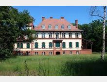 Historisches Herrenhaus mit symmetrischer Fassade, roten Dachziegeln, grünen Fensterläden und einem Sockel aus rotem Mauerwerk, umgeben von einer Wiese und Bäumen unter klarem, blauem Himmel.