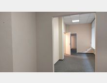 Leerer Büroraum mit hellbeigen Wänden, grauem Teppichboden und einer weißen Zwischentür, die in einen weiteren Raum führt. Rechts im Bild befindet sich ein großes Fenster mit vertikalen Lamellen-Jalousien und einer Steckdose an der Wand.