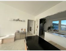 Heller Wohnraum mit offener Küche, weiß gestrichenen Wänden und großen Fenstern. Im Vordergrund steht ein schwarzer Tisch, an der Wand befinden sich ein schwebendes Regal mit Gläsern und Geschirr sowie ein Sideboard, im Hintergrund sind Pflanzen und eine Küche mit Sicht ins Freie sichtbar.