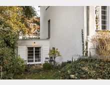 Außenansicht eines weißen Wohnhauses mit einer Fensterfront, die von geöffneten Fensterläden umrahmt ist. Im Vordergrund ist ein Garten mit Rasenfläche, Büschen und einer rankenden Pflanze an der Hauswand zu sehen.