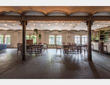 Ein geräumiger, rustikaler Innenraum mit Holzboden, sichtbaren Säulen und einer gewölbten Decke. Mehrere Holztische und -stühle stehen lose arrangiert, umgeben von großen Fenstern, die viel Tageslicht hereinlassen; im Hintergrund sind Pflanzen, ein Klavier und eine Bar mit Hockern zu sehen.