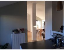 Ein offener Wohnbereich mit Blick von der Küche in einen Essbereich. Im Vordergrund eine dunkle Küchenarbeitsplatte mit Küchengeräten, im Hintergrund ein Esstisch aus Holz, Stühle und ein Fenster, das Tageslicht hereinlässt. Ein Heizkörper mit Dekoration in Form von Trockenblumen steht vor einer grünen Wand.