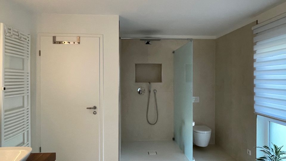 Modernes Badezimmer mit einer bodengleichen Dusche, einer halbtransparenten Glasabtrennung, einem weißen Wand-WC und einem großen Fenster mit Rollos rechts. Links sind ein Heizkörper, eine Tür und ein Waschtisch mit Holzelementen sichtbar.