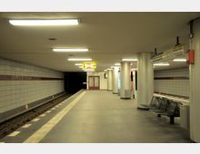 Eine U-Bahn-Station mit dem Namen "Kleistpark", gekennzeichnet durch helle rechteckige Fliesenwände mit einem roten Streifen. Der Bahnsteig ist beleuchtet, enthält Sitzbänke, Informationsschilder, Mülleimer und mehrere Säulen, die den Raum strukturieren, mit Gleisen auf der linken Seite.