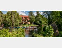 Eine Flusslandschaft mit einem Holzhaus mit roten Dachziegeln im Hintergrund, umgeben von Bäumen und grüner Vegetation. Im Vordergrund eine Gruppe von Menschen auf einem Boot, weitere kleine Boote sind auf dem Wasser zu sehen, und rechts steht ein Strohhaufen auf einer Wiese.