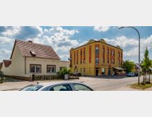 Straßenszene mit einem kleinen Einfamilienhaus links und einem gelben, dreigeschossigen Gebäude mit roter Fassade rechts, in dem sich eine Bäckerei und ein Café befinden. Im Vordergrund ist ein geparktes Auto zu sehen, und die Straße wird von einem Laternenmast gesäumt.