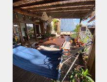 Überdachte Dachterrasse mit Holzboden, Holzlattenpergola und Pflanzen in Töpfen. Einrichtung umfasst eine Hängematte, Holzmöbel und ein Ecksofa, umgeben von begrünten Elementen und großem Ausblick auf städtische Dächer.