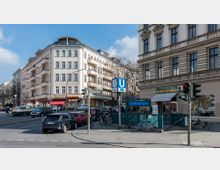 Straßenszene in einer Stadt mit Wohn- und Geschäftshäusern im Altbaustil, einem Eingang zur U-Bahn-Station "Kleistpark" der Linie U7 sowie Fahrrädern und parkenden Autos. Im Vordergrund sind Ampeln, Straßenschilder und ein Blumenladen sichtbar.