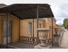 Gelbes historisches Gebäude mit rustikaler Veranda, flankiert von dekorativen Steinvasen auf einem Geländer. Rechts verläuft ein sandiger Weg entlang einer Arkade und an Bäumen vorbei.