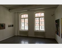 Ein heller Galerieraum mit weiß gestrichenen Wänden, zwei hohen Fenstern mit Blick auf ein gegenüberliegendes Backsteingebäude, Wandheizkörpern unter den Fenstern und mehreren Gemälden an den Wänden.