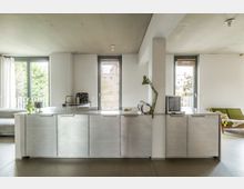 Moderne, minimalistische Küche mit einer Arbeitsplatte aus Edelstahl, großen Schubladenelementen und integriertem Kochfeld. Im Hintergrund sind bodentiefe Fenster mit Blick ins Freie, Vorhänge und eine Sitzecke mit grünem Sofa zu sehen.