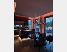 Moderne Küchen- und Esszimmerkombination mit dunklen Möbeln und warmweißer indirekter Beleuchtung. Große Fenster und Schiebetüren bieten Blick auf eine Terrasse mit Sitzmöbeln und einen urbanen Außenbereich.